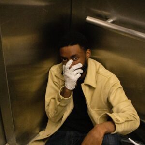 Rapper Kaiser Unique sitting on the floor of an elevator with a white gloved hand over his face