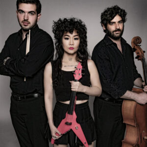 a man holding drumsticks stands and a man with a cello stand on each side of a woman holding a pink electric violin