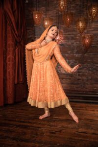 A dancer dressed in peach-colored Indian clothing stands with one toe pointed and one hand outstretched in front of a rustic wall with lanterns and a red curtain.