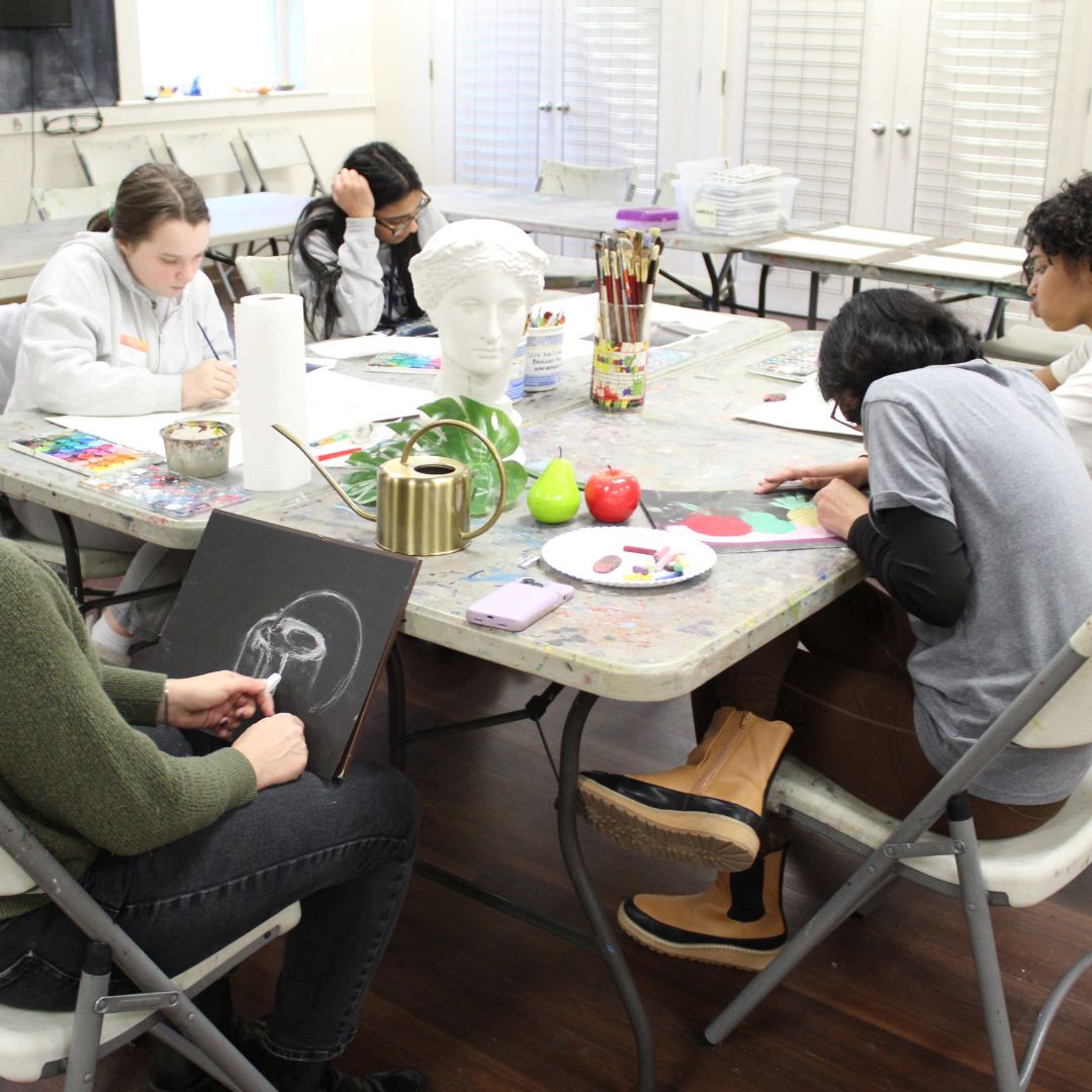 group of 4 students around a table with their teacher working on various paintings
